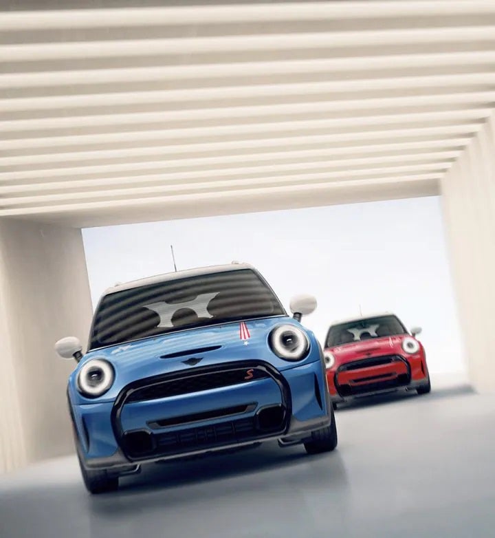 Front views of two MINI 20 Years Edition vehicles, one in Island Blue and one in Chili Red, underneath a square beige entranceway with stripes overhead and a clear sky in the background. | MINI of Sterling in Sterling VA