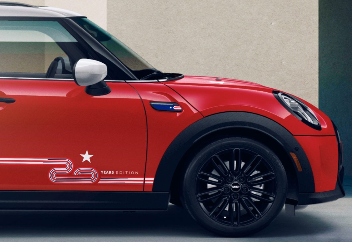 Side view of a Chili Red MINI 20 Years Edition Hardtop 4 Door Cooper S parked on a grey surface with a beige building in the background. | MINI of Sterling in Sterling VA