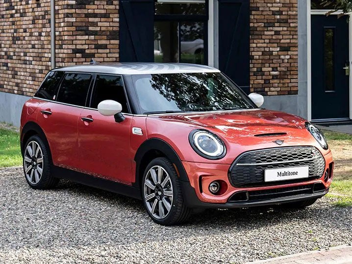 Three-quarters front view of a MINI Clubman Multitone Edition in Coral Red parked in front of a brown brick building.