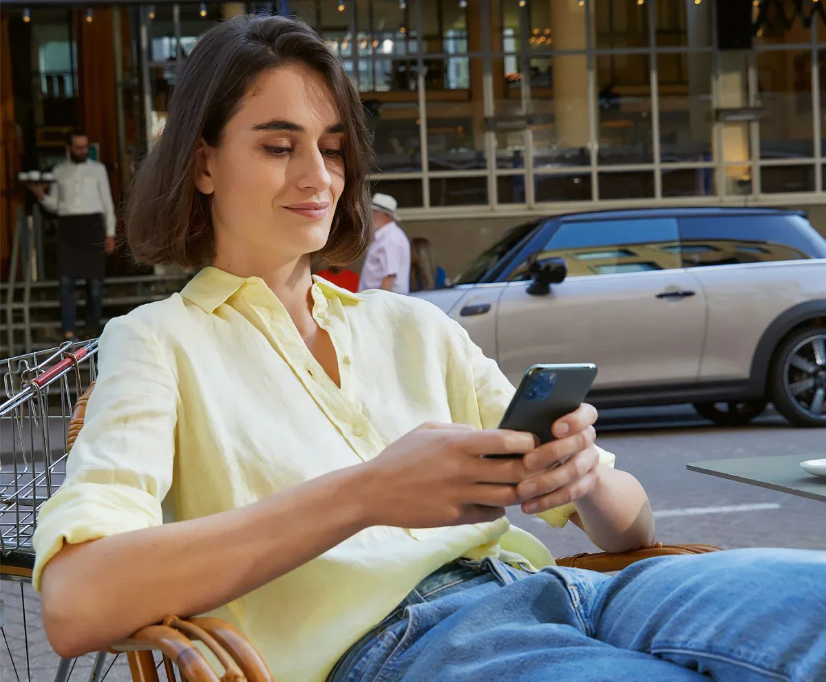 Female wearing yellow collared button-down shirt and jeans, sitting in a chair and holding a smartphone next to a street with a grey MINI vehicle parked in the background. | MINI of Sterling in Sterling VA