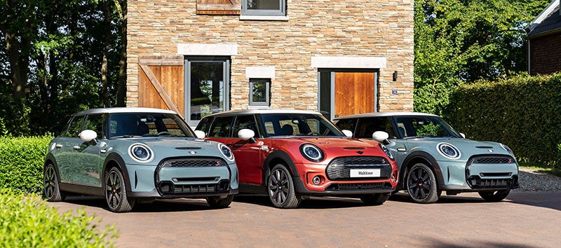 Three MINI Multitone Edition vehicles, two in Sage Green and one in Coral Red, parked side-by-side on a brick surface in front of a brown brick building with greenery surrounding it.